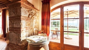 Ein rustikaler Raum mit einer Steinwand, einem Glastisch mit Büchern und Blick auf eine Außenpatio mit Stühlen und Sonnenschirmen.