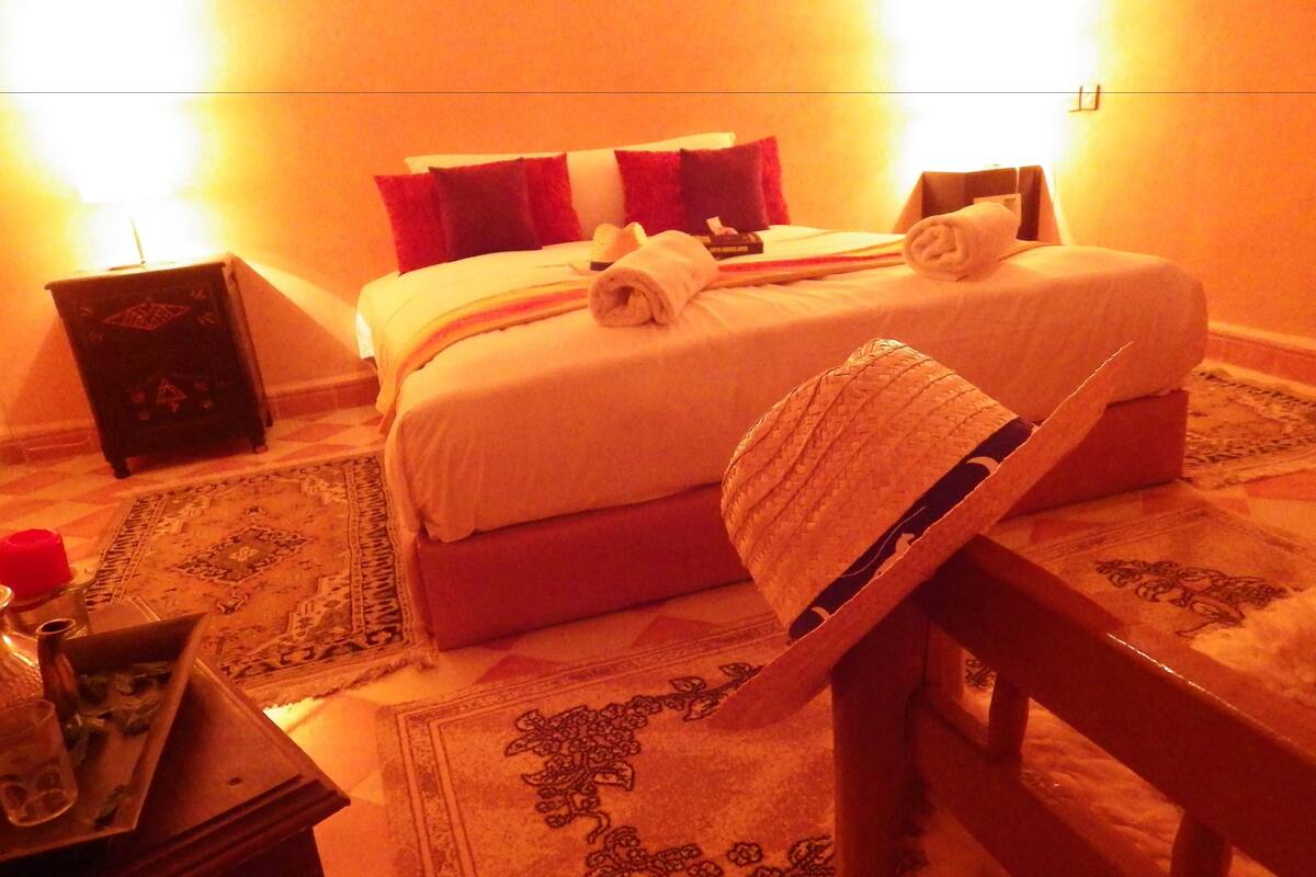 A hotel room with a bed, a wooden chair, a small table with a glass and a book, and a decorative rug.