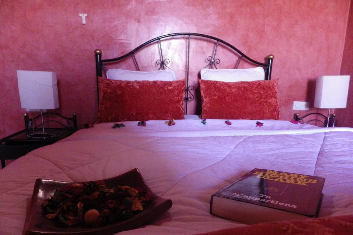 A bed with a white comforter and red pillows, a bedside table with a lamp, and a book titled "The Art of Cooking" on the bed.