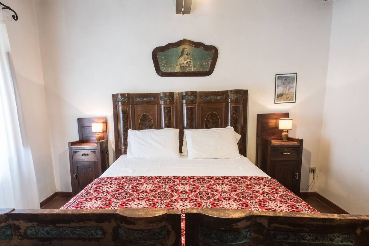 A bedroom with a large bed, two bedside tables, a nightstand, a decorative mirror, and a wall hanging.