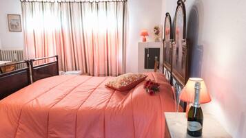 A bedroom with a bed, a nightstand with a bottle of wine, a lamp, and a window with curtains.