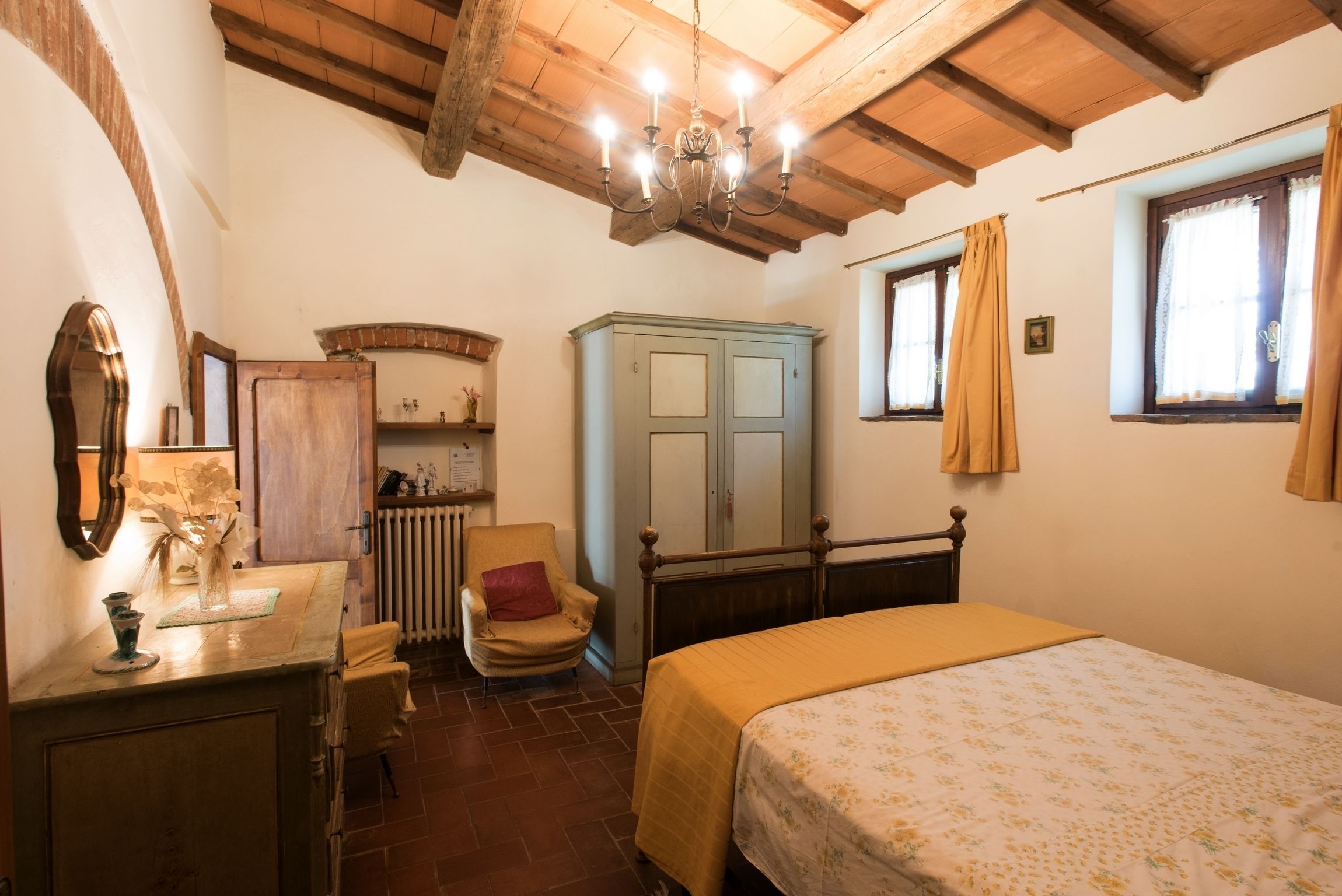A bedroom with a wooden ceiling, a bed with a floral bedspread, a chair, a wardrobe, and a radiator.