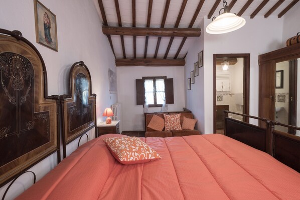 Un dormitorio con una cama grande, cabecera de madera, una mesita de noche, una lámpara y una ventana con contraventanas.