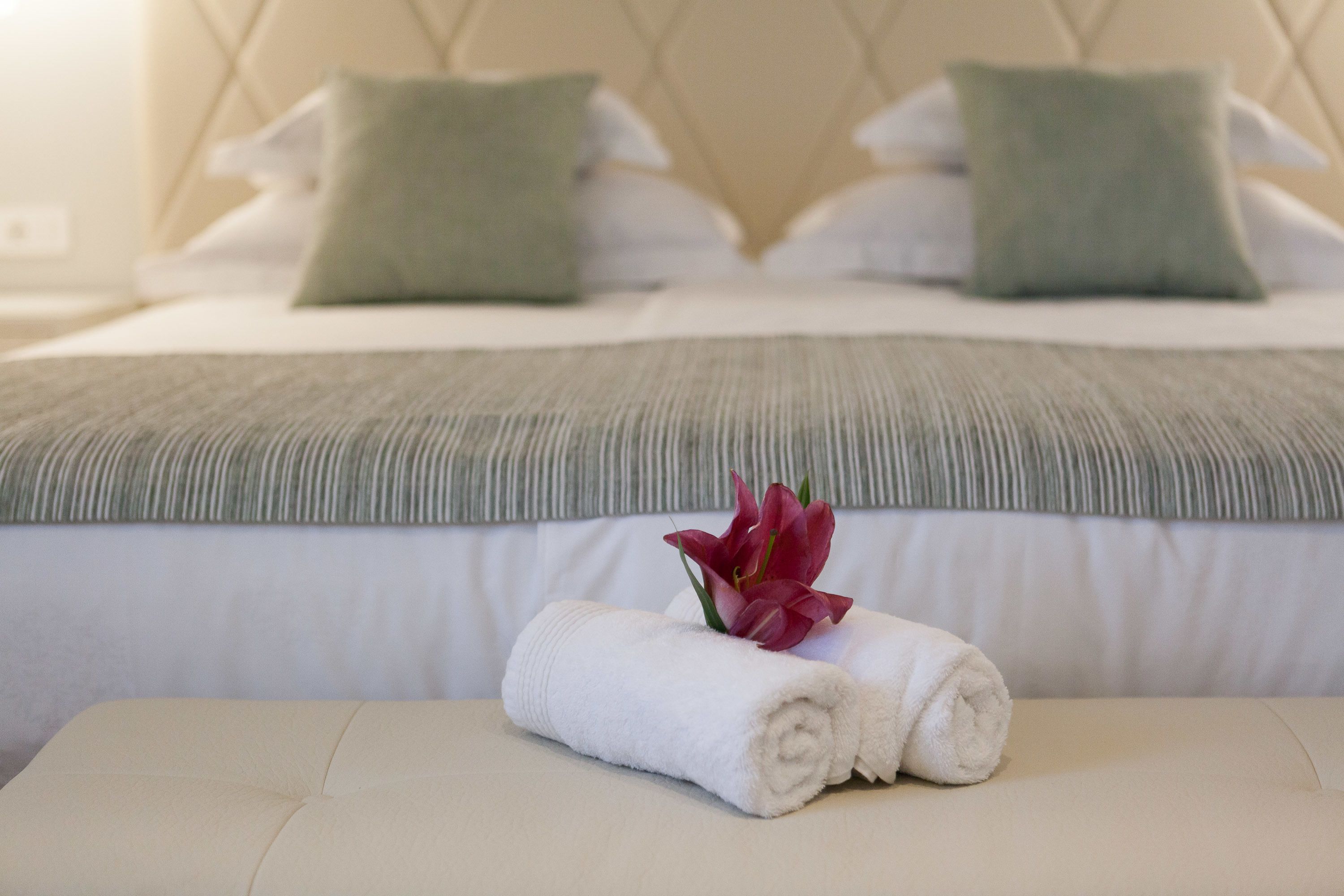 A neatly made bed with a green striped blanket, two pillows, and a rolled white towel with a red flower on a bedside table.