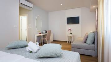 Quarto de hotel moderno com uma cama, um sofá, uma escrivaninha com cadeira, uma mesinha com planta e uma TV na parede.