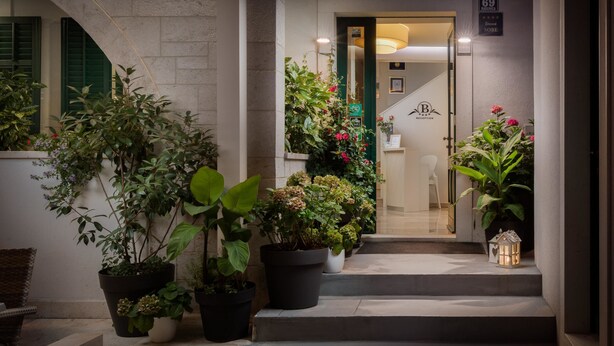 A hotel entrance with potted plants, a bench, and a staircase leading to the interior.