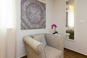Una poltrona dai toni neutri con un cuscino, un'opera d'arte in mosaico, un'orchidea in vaso e uno specchio che riflette un bagno.