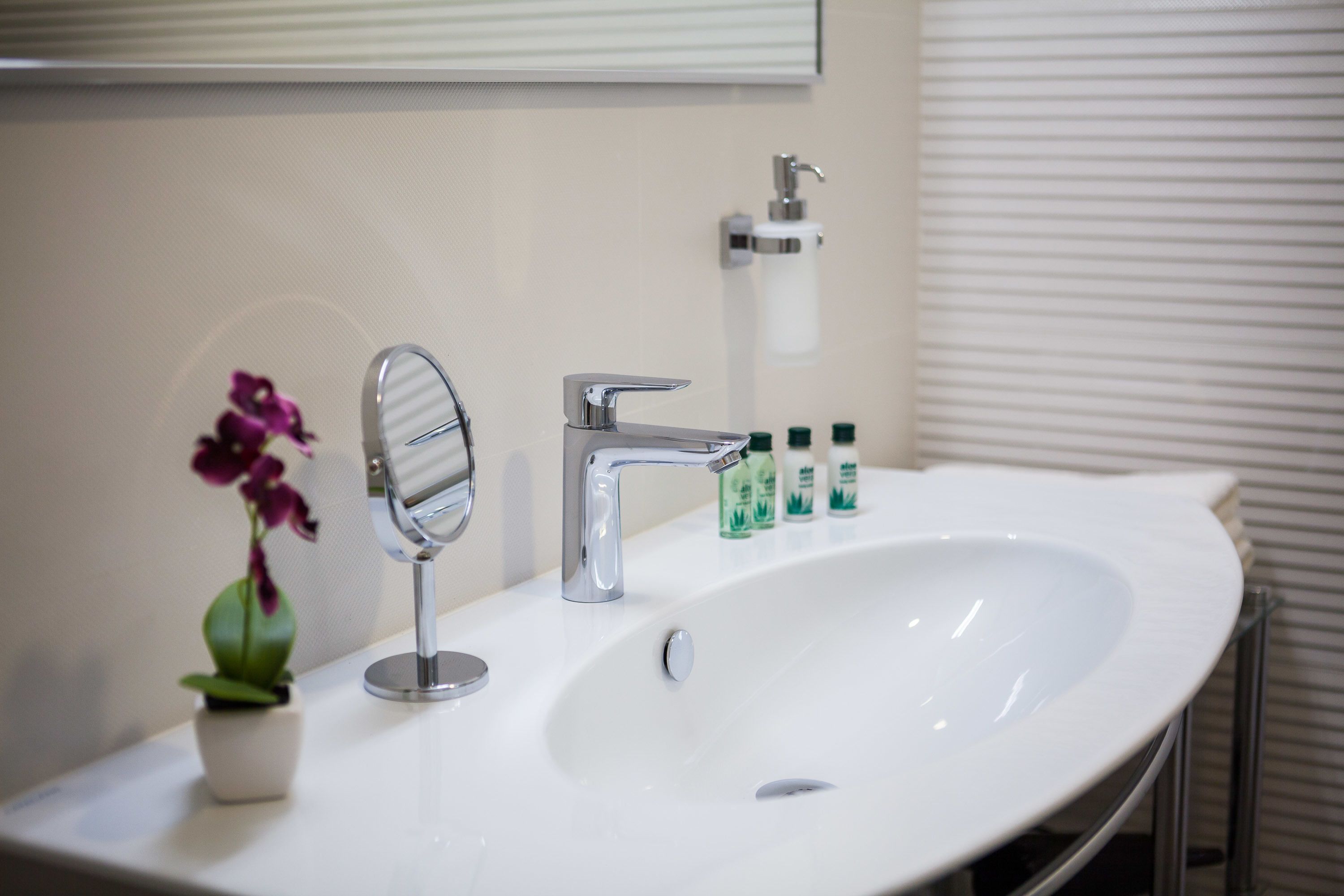 A bathroom with a white sink, a mirror, a faucet, and a small potted orchid.