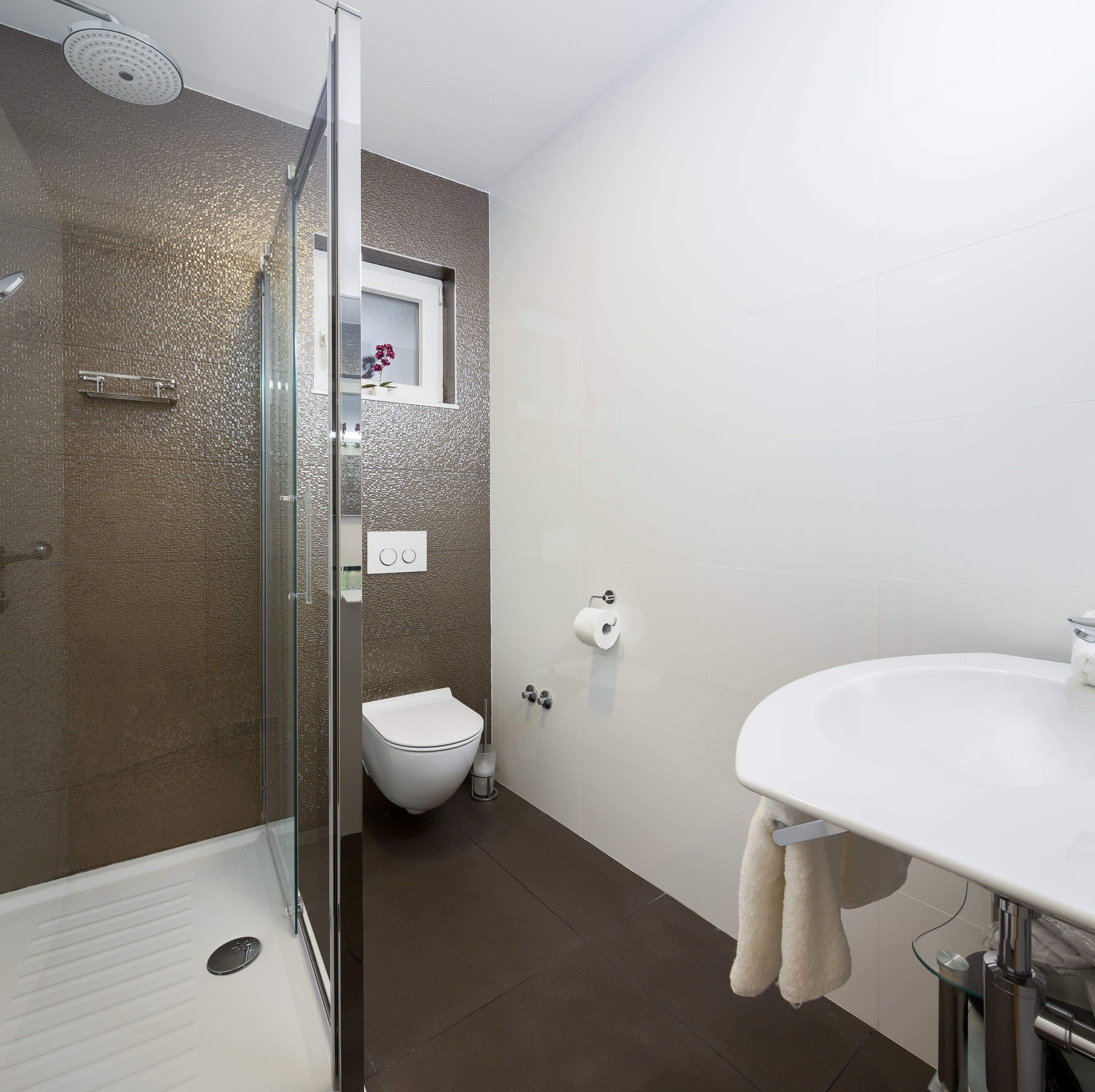 A modern bathroom with a glass shower enclosure, a toilet, and a sink with a towel hanging on the wall.