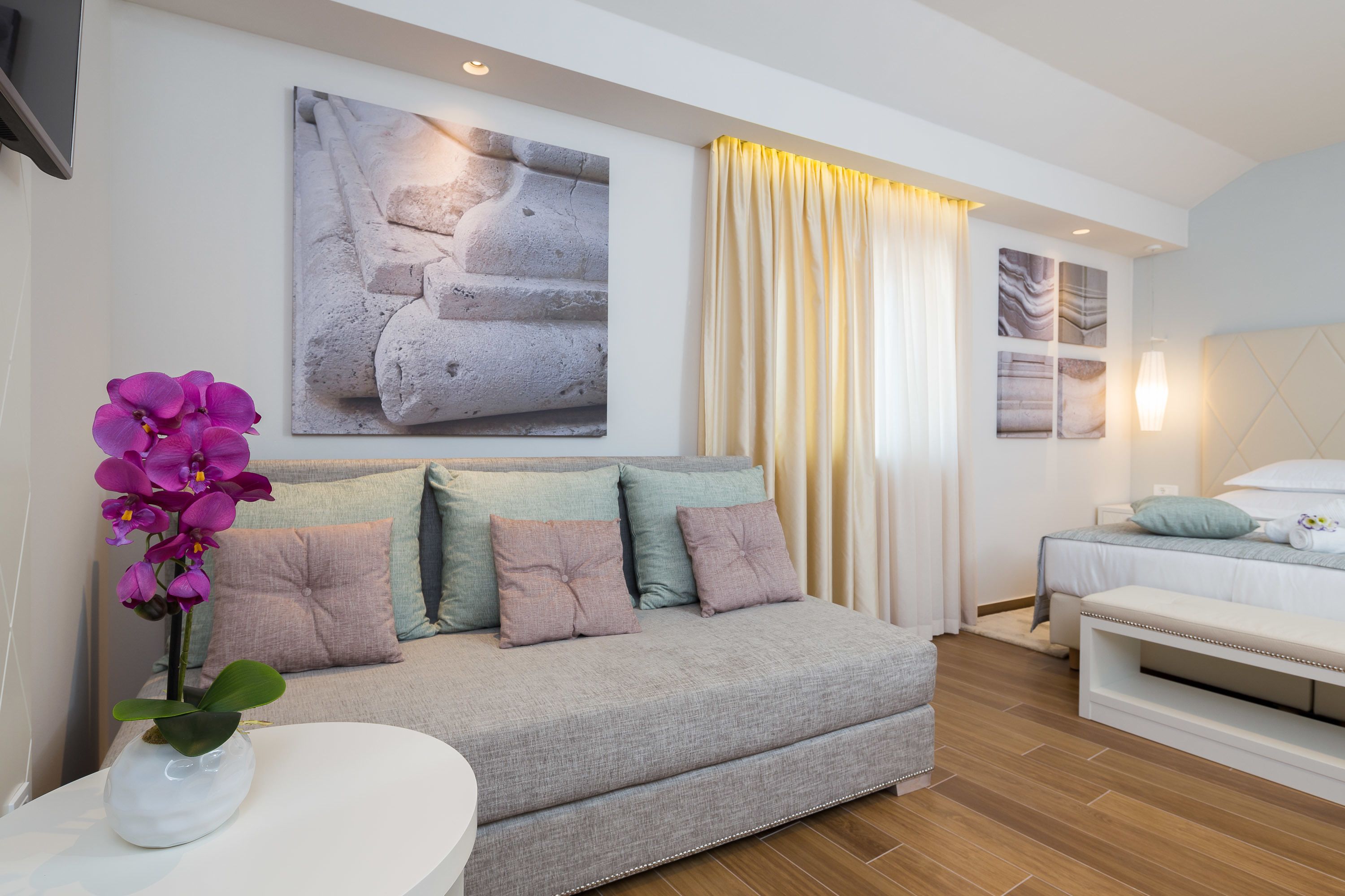A modern hotel room with a grey sofa, a bedside table with a vase of purple orchids, a bed with white bedding, and artwork on the walls.