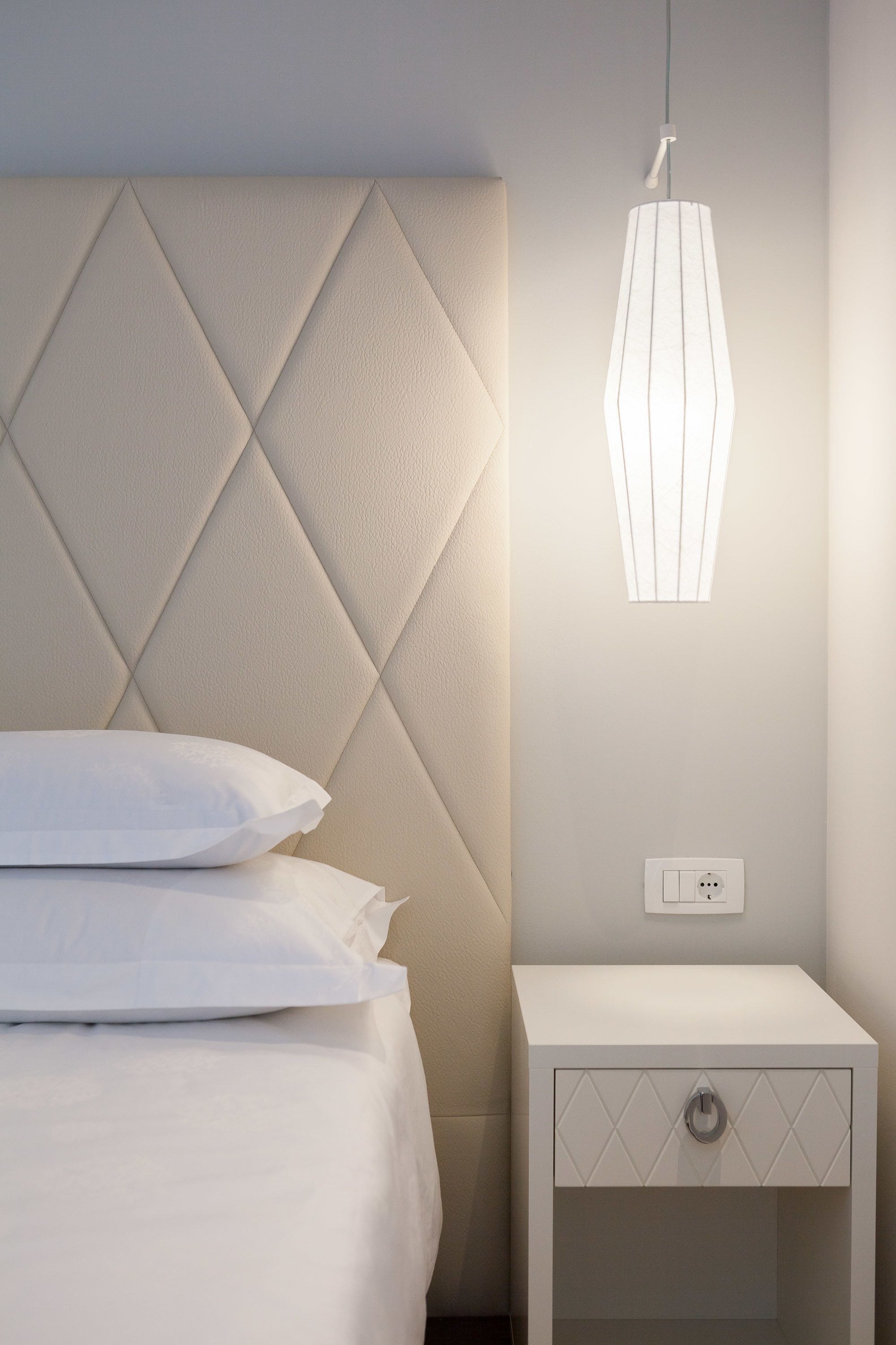 A neatly made bed with white linens and pillows, a modern headboard with a diamond pattern, a bedside table with a drawer, and a hanging pendant light.