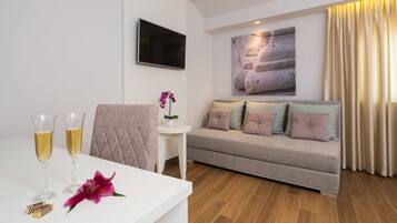 A modern living room with a sofa, a flat-screen TV mounted on the wall, a small table with two glasses of champagne, and a decorative flower.