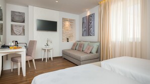 A hotel room with a bed, a sofa, a desk with a chair, a TV, and artwork on the walls.