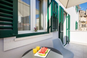 Una mesa con una fuente de sandía en rodajas y dos vasos de jugo de naranja, una silla y un ventanal con contraventanas verdes.