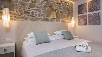 Una cama bien hecha con sábanas blancas y almohadas verdes, una pared de piedra con una pequeña estatua, y dos lámparas fijadas a la pared.