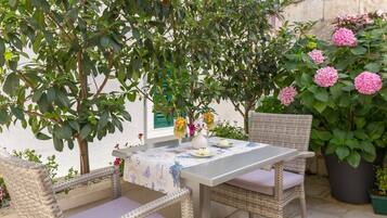 Een terras met rieten stoelen en een tafel voor twee personen, omgeven door weelderige begroeiing en roze bloemen.