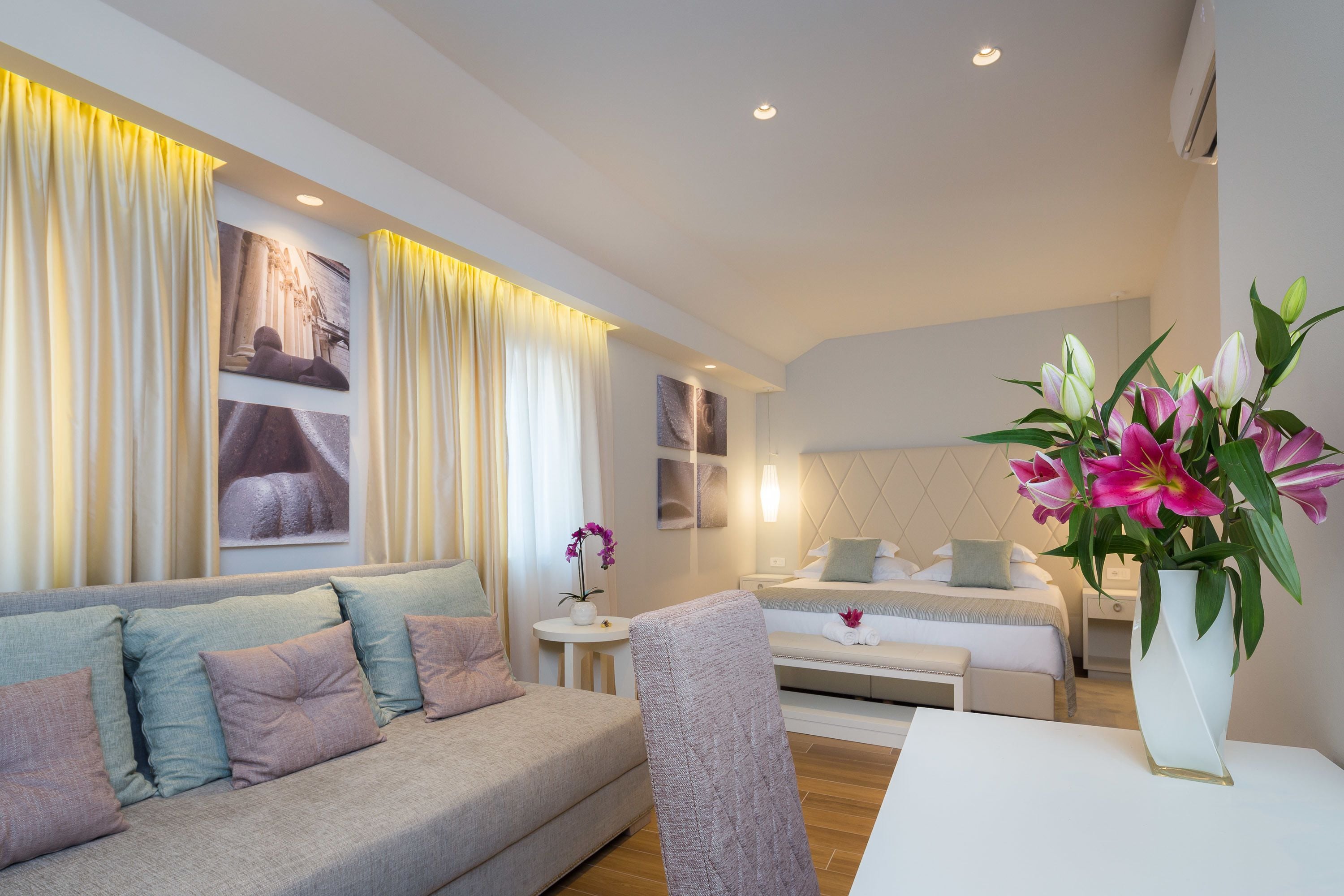 Una habitación de hotel moderna con sofá, cama, una mesita con un florero y una obra de arte colgada en la pared.