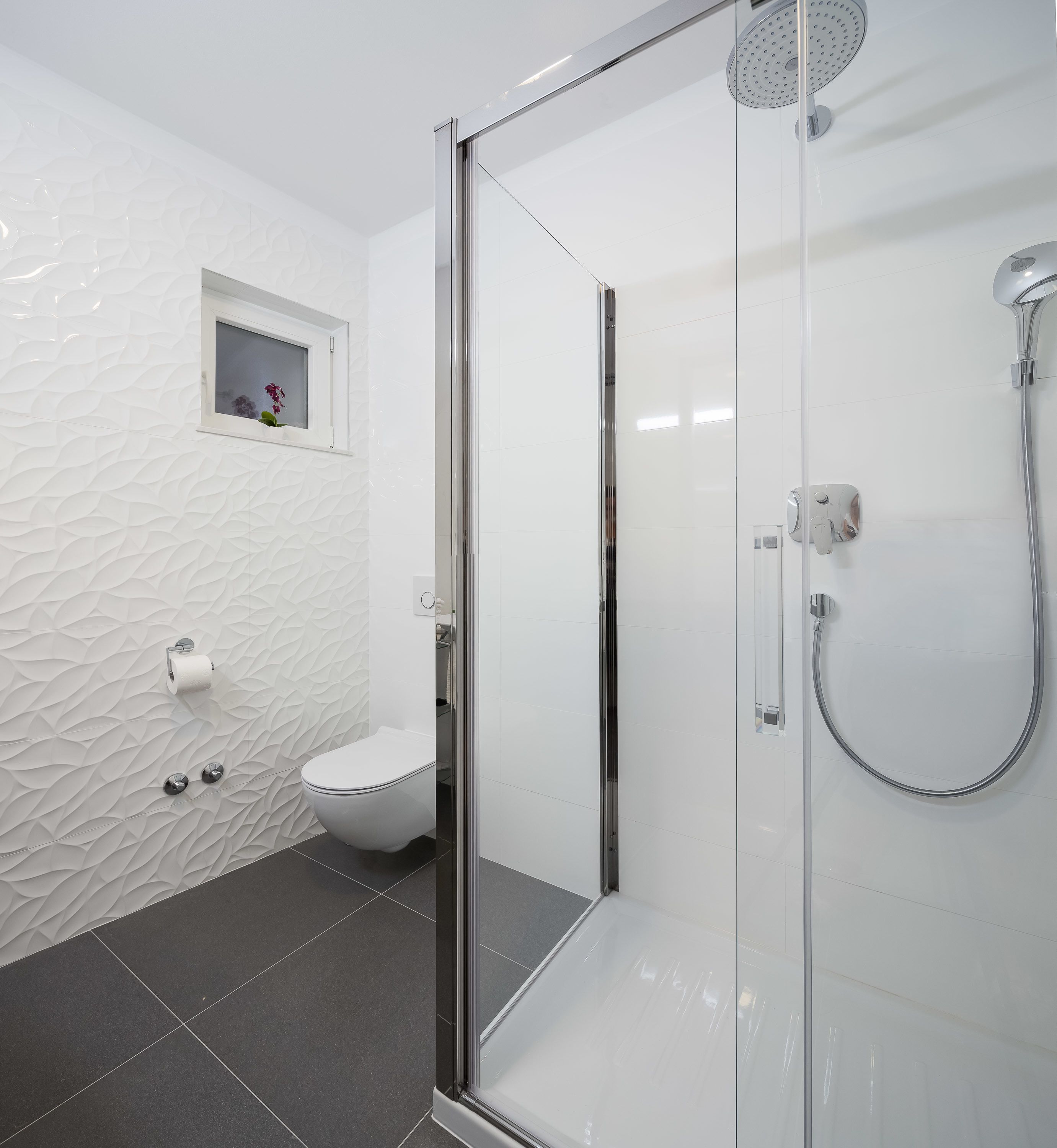 Ein modernes Badezimmer mit Duschkabine aus Glas, Toilette und kleinem Fenster mit Blumen.