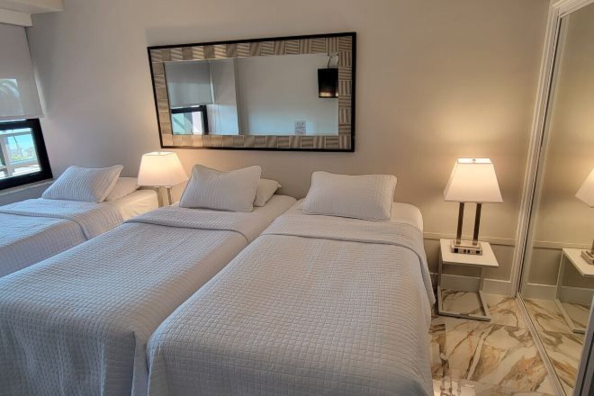 Habitación de hotel con dos camas, un espejo grande, lámparas de noche y suelo de mármol.