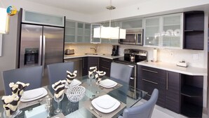 A modern kitchen with a glass dining table, stainless steel appliances, and dark cabinetry. - Deluxe Beach Resort - HORA RENTALS (Miami Beach)