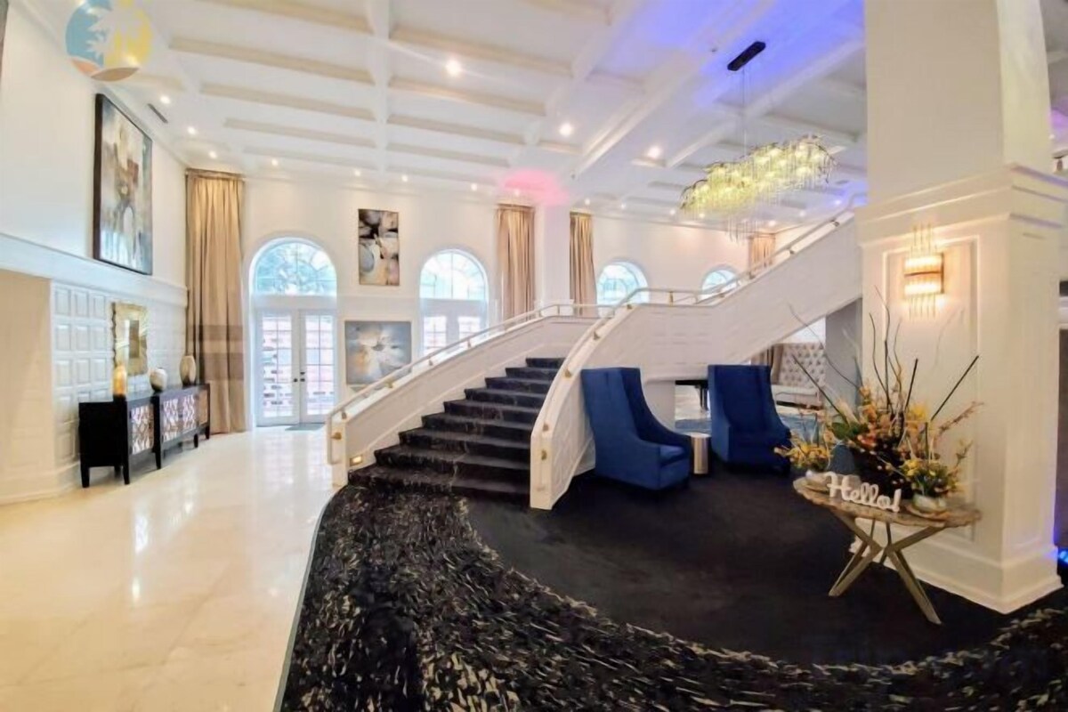 A grand staircase leading to an upper level, a reception area with a desk, and a decorative plant arrangement.