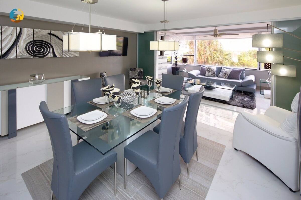 A modern dining area with a glass table and blue chairs, a sofa, and a view of the outdoors.