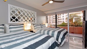 A hotel room with two beds, a ceiling fan, a decorative wall panel, and a balcony with a view of other buildings and greenery. - Deluxe Beach Resort - HORA RENTALS (Miami Beach)