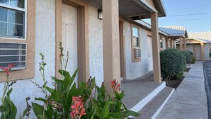A row of single-story buildings with a beige exterior, a covered entrance, and a small garden area with pink flowers. - Desert Inn (Van Horn)