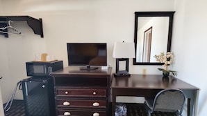 A hotel room with a desk, a television, a lamp, a trash bin, a chair, a microwave, and a small plant. - Desert Inn (Van Horn)