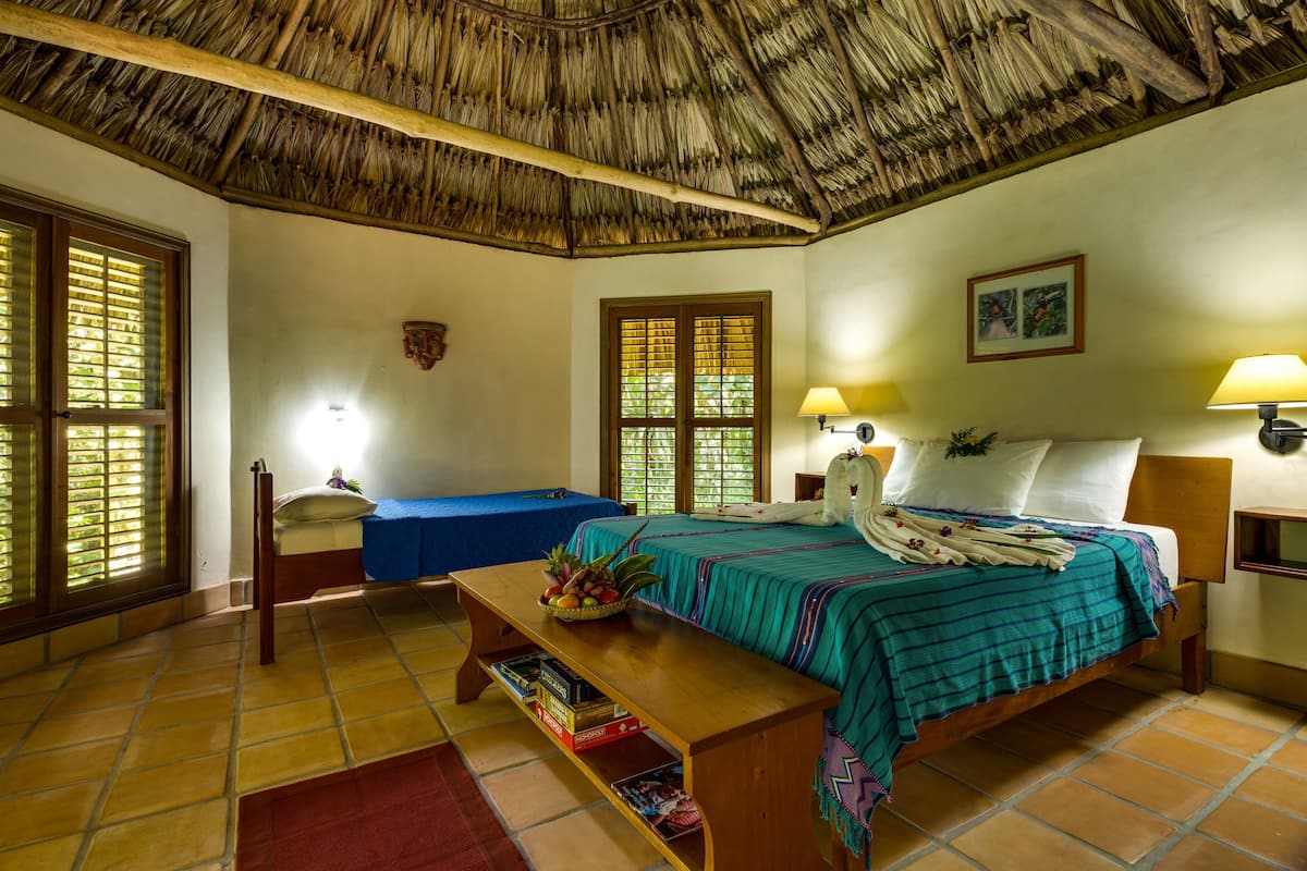 A room with a bed, a bench, a table with books, a window with shutters, and a thatched ceiling.