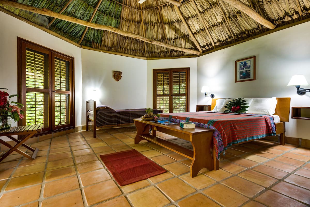 A room with a bed, a wooden table, a red rug, and a thatched roof ceiling.