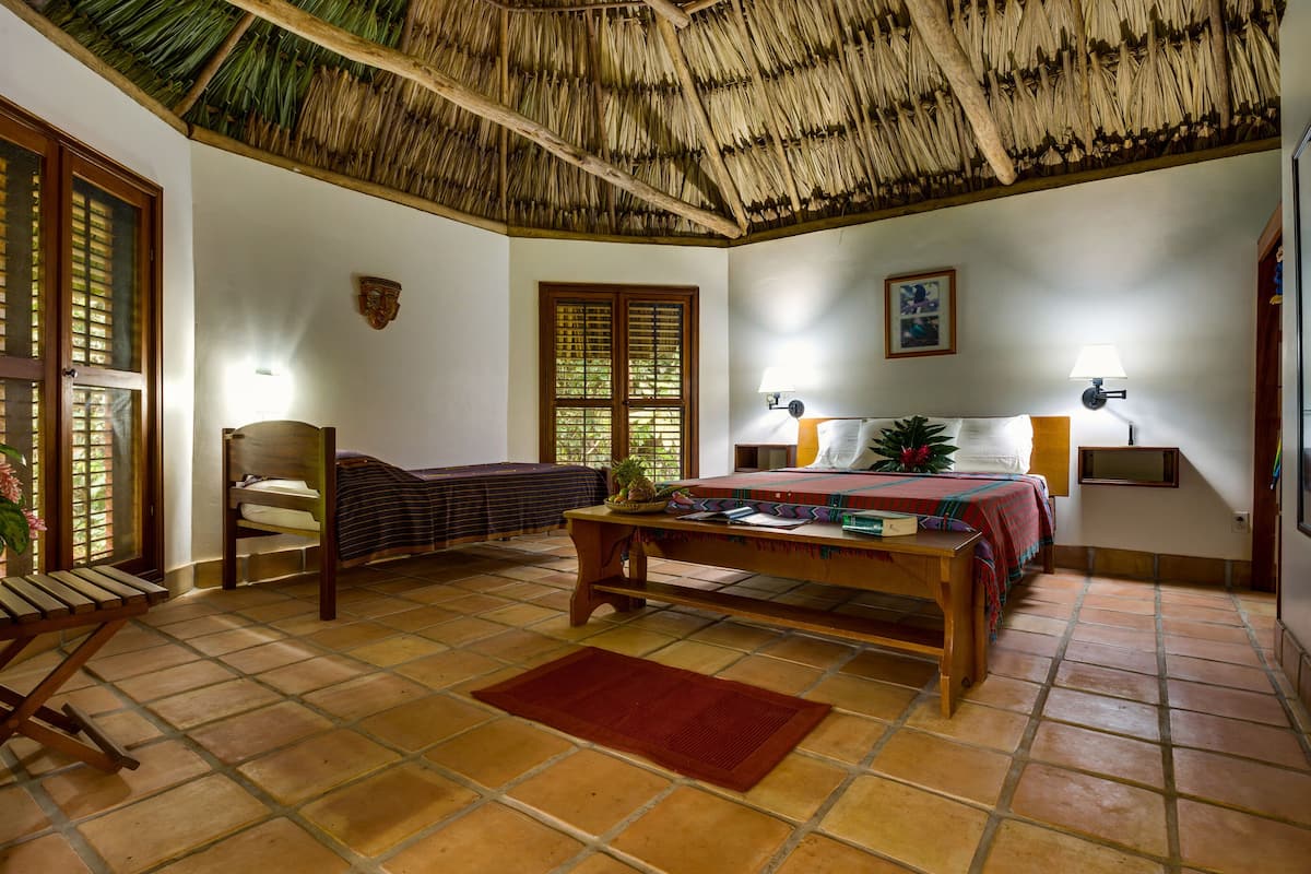 A room with a bed, a wooden table, a chair, a window with shutters, a wall-mounted lamp, and a thatched roof ceiling.