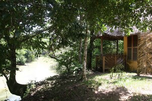 Una cabaña rústica con techo de paja, rodeada de densa vegetación y una masa de agua tranquila.