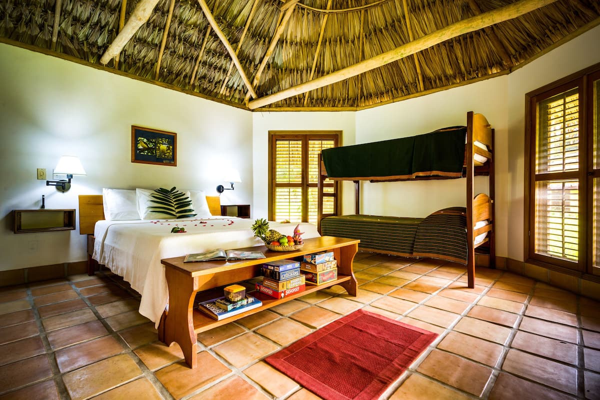 A room with a bed, a wooden bench, a table with books, a thatched ceiling, and a window with blinds.