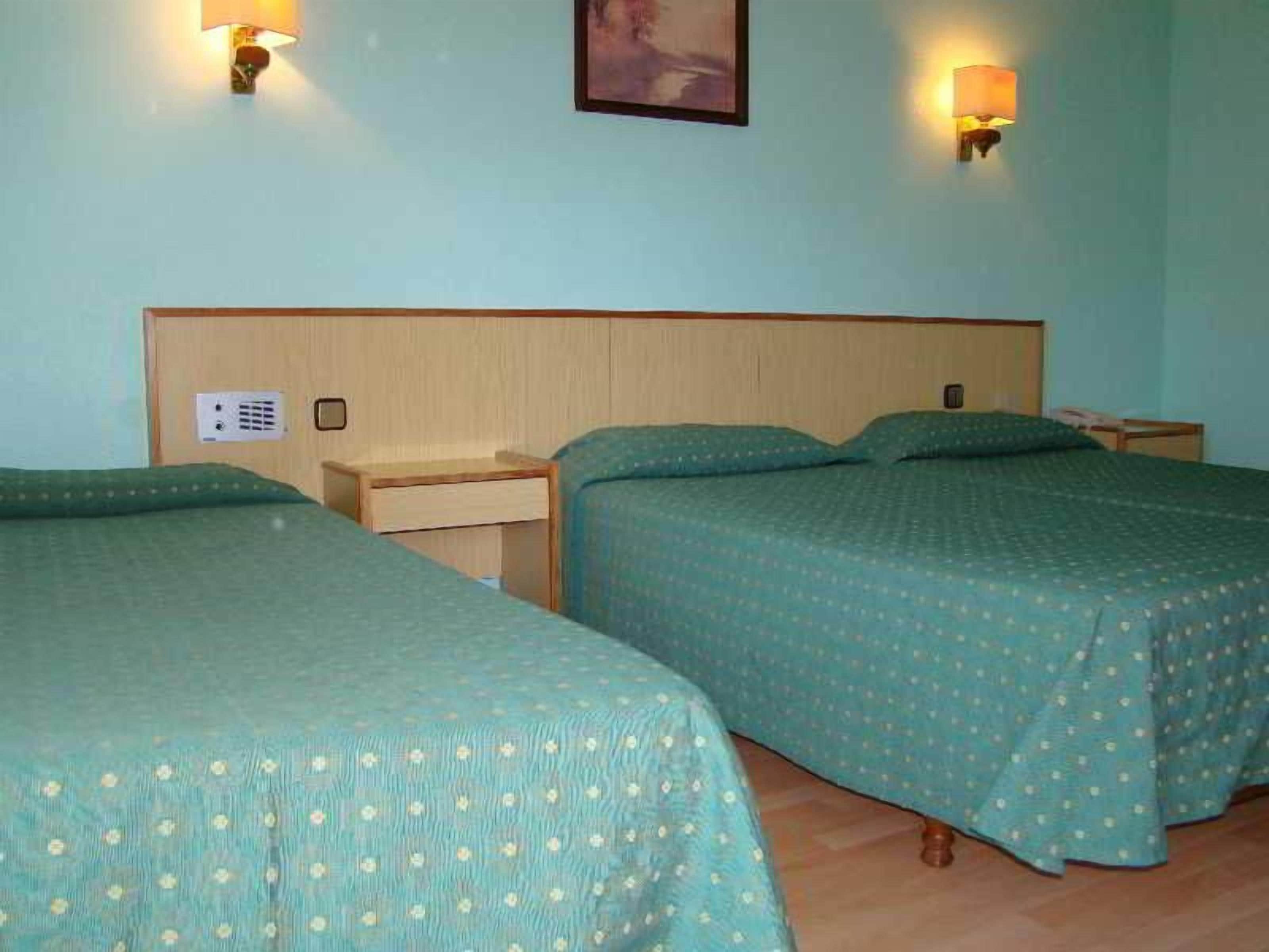 Quarto de hotel com duas camas de solteiro, cabeceira de madeira, mesas de cabeceira e luminárias fixadas na parede.