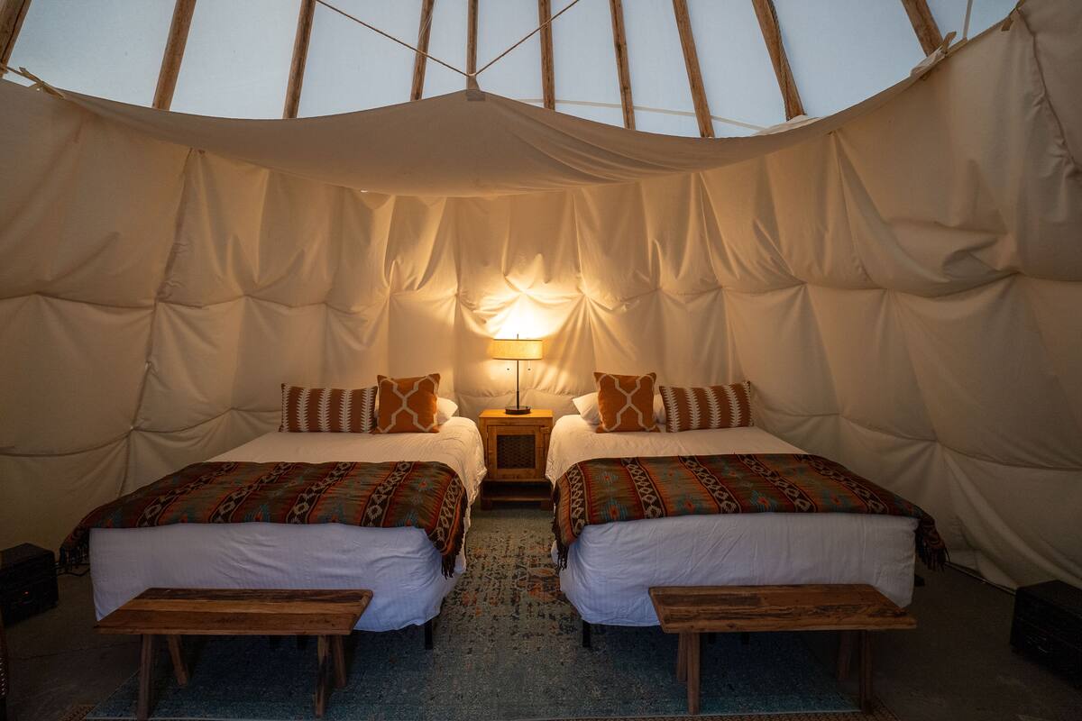 A tented accommodation with two beds, a central nightstand with a lamp, and a blue carpet.