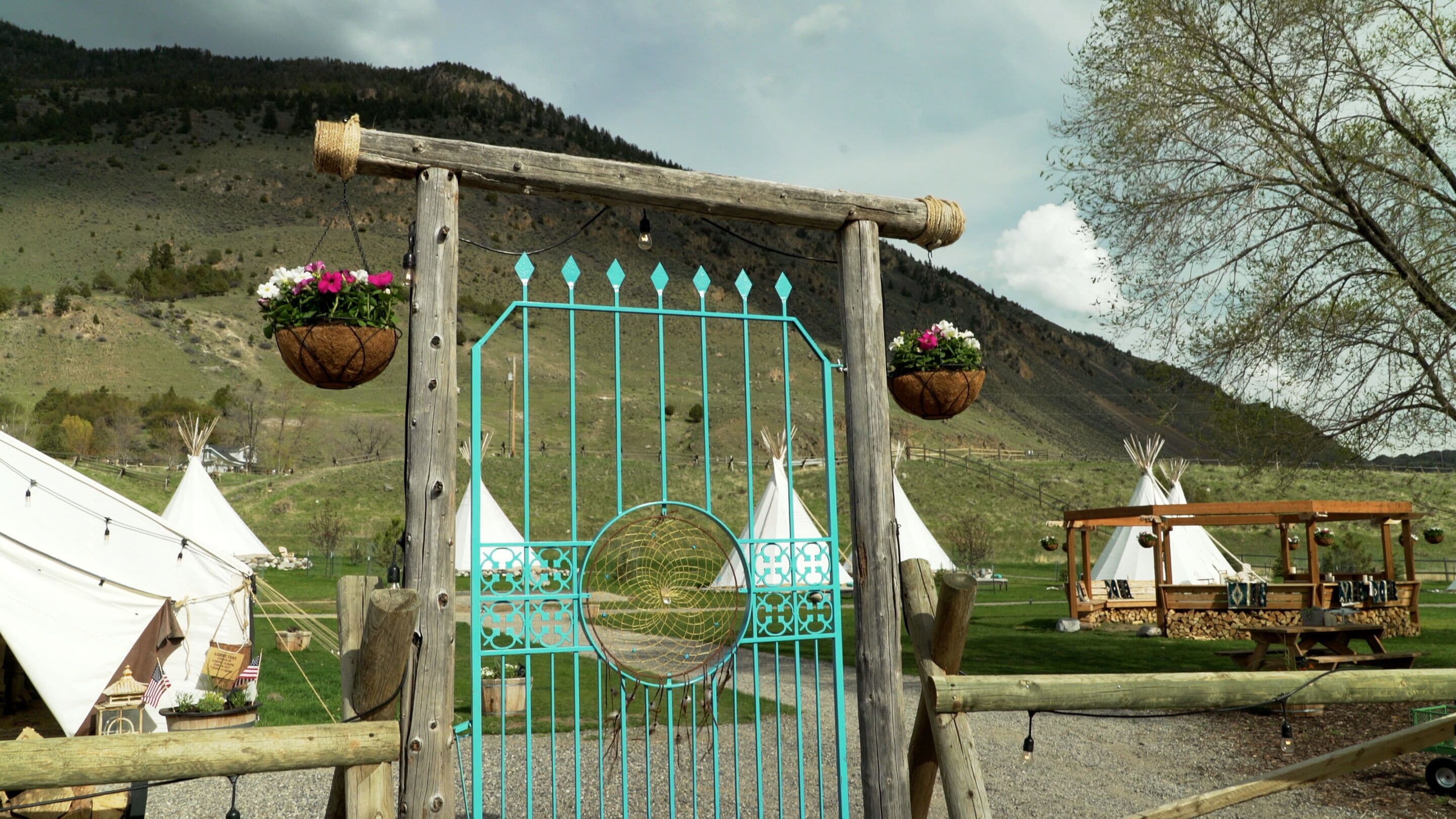A wooden gate with a turquoise metal design, surrounded by white tents and a wooden structure with hanging flower baskets.