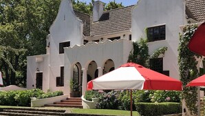 Zona de piscina con sillas de descanso y sombrillas a rayas rojas y blancas, rodeada por un edificio blanco con arcos y techo de tejas.