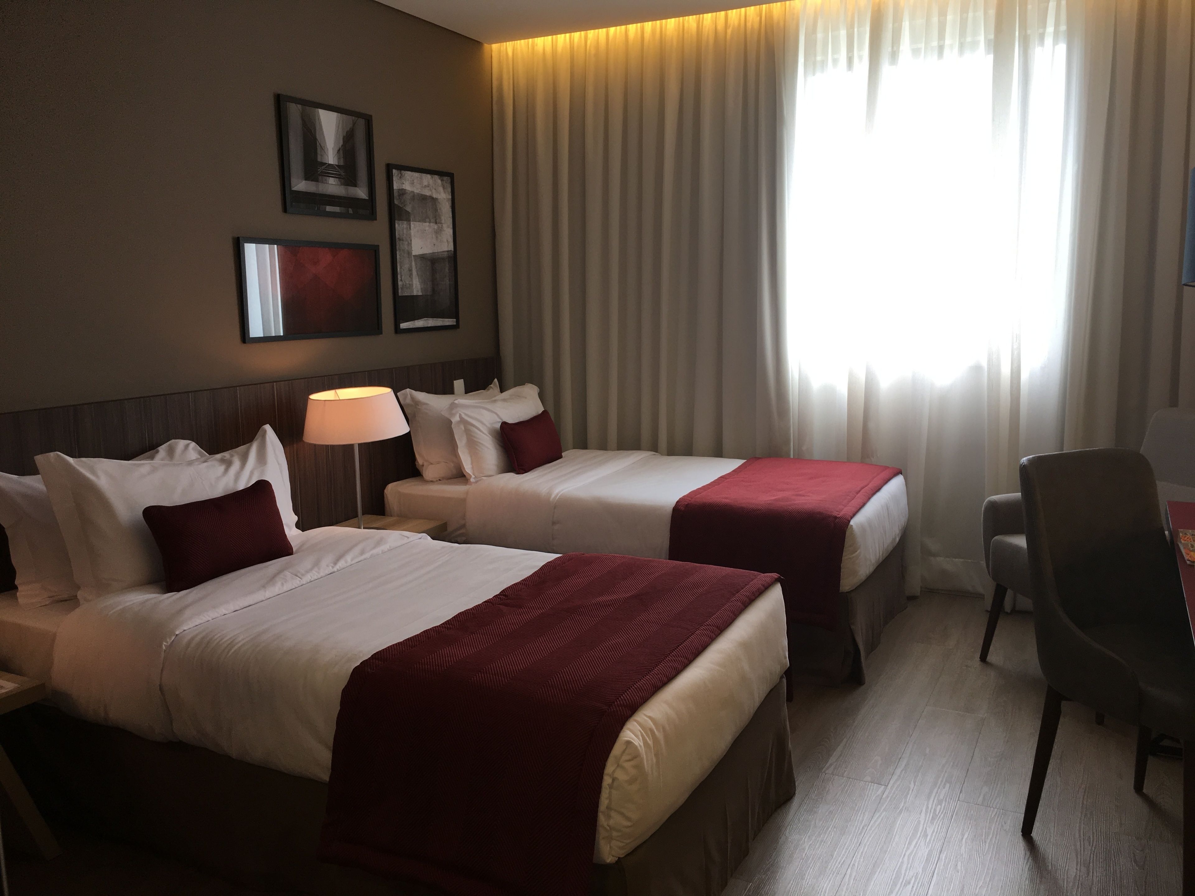 A hotel room with two beds, a desk, and a chair. There are curtains on the window and framed pictures on the wall.