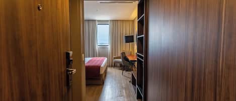 A hotel room with a wooden floor, a bed with a red blanket, a desk with a chair, a television on the wall, and a window with curtains.