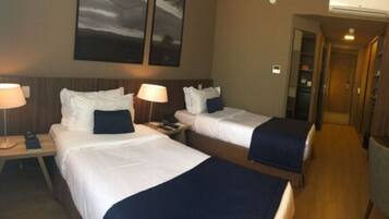 A hotel room with two beds, each with a pillow and a blanket. There are bedside tables with lamps and a wall-mounted air conditioning unit.