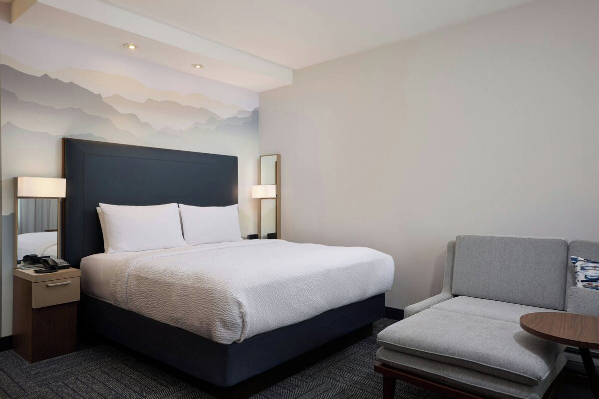 A hotel room with a large bed, a bedside table with a lamp, a chair, and a small table.