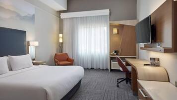 Habitación de hotel con una cama grande, un escritorio, una silla, un televisor y una ventana con cortinas.