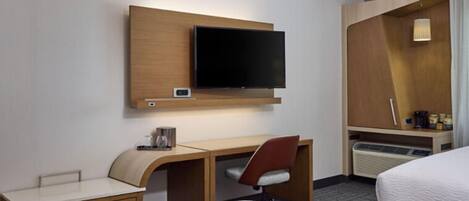 A hotel room with a bed, a desk with a chair, a television mounted on the wall, and a small table with a lamp.