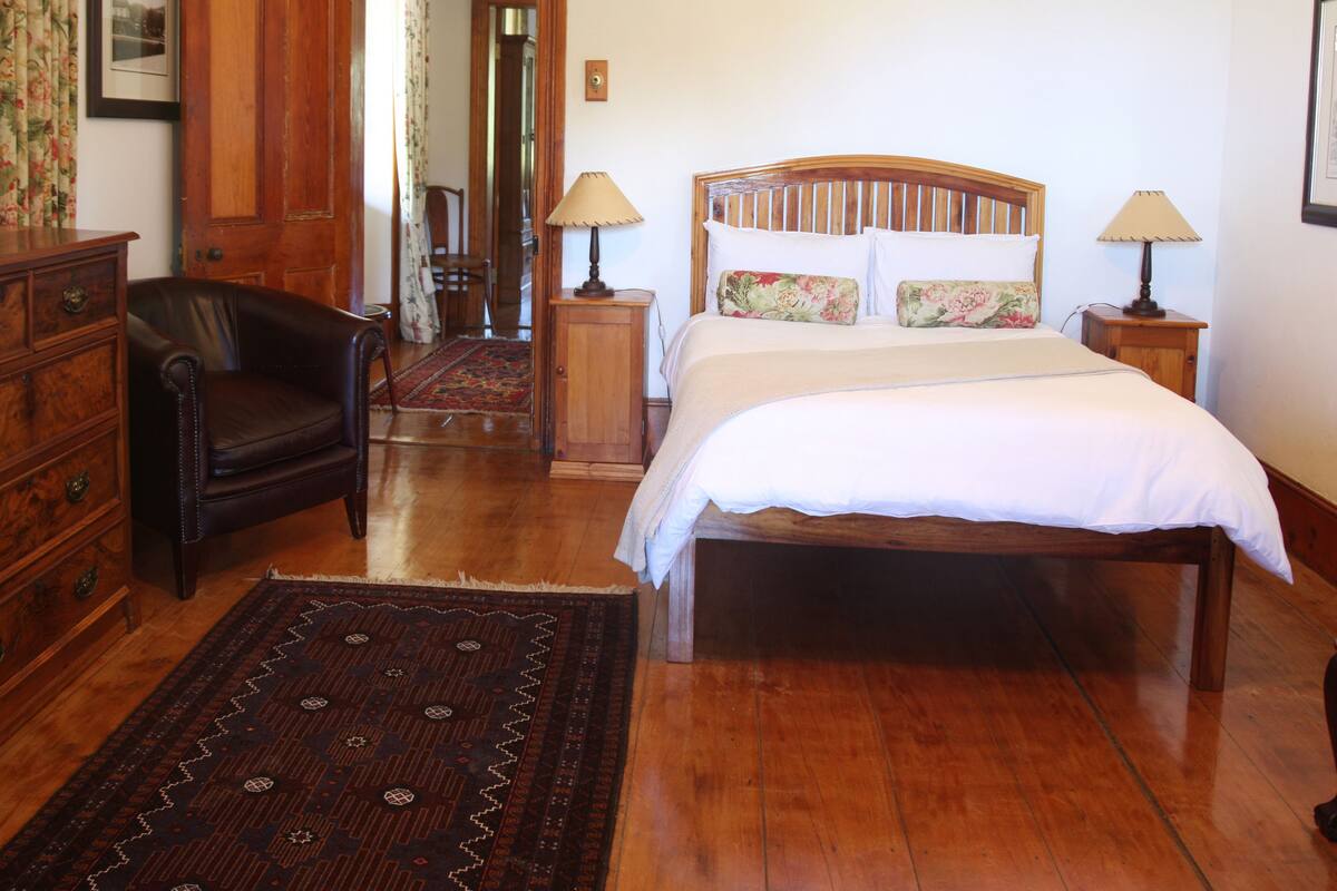 Un dormitorio con cama de madera, dos mesitas de noche con lámparas, un aparador de madera, una silla y una alfombra estampada.