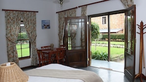Une chambre à coucher comprenant un lit, une table de chevet en bois, une table avec des chaises, une fenêtre avec des rideaux et une porte donnant sur un jardin.