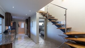A modern kitchen with a granite countertop, a stainless steel sink, and wooden cabinets. There is a staircase with wooden steps and a glass railing leading to another level. - Dockside Guest House (Port Alfred)