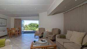 A spacious living room with a glass coffee table, beige sofas, and a dining area with wooden chairs. - Dockside Guest House (Port Alfred)