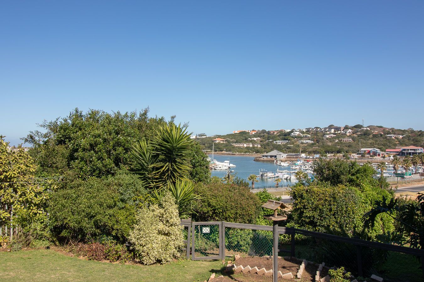 Vista costera con un puerto deportivo, casas en una ladera y una puerta con un cartel.