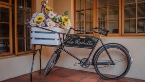 A bicycle with a basket and flowers, parked on a tiled floor next to a building with windows. - Guesthouse LaRachelle (Swellendam)
